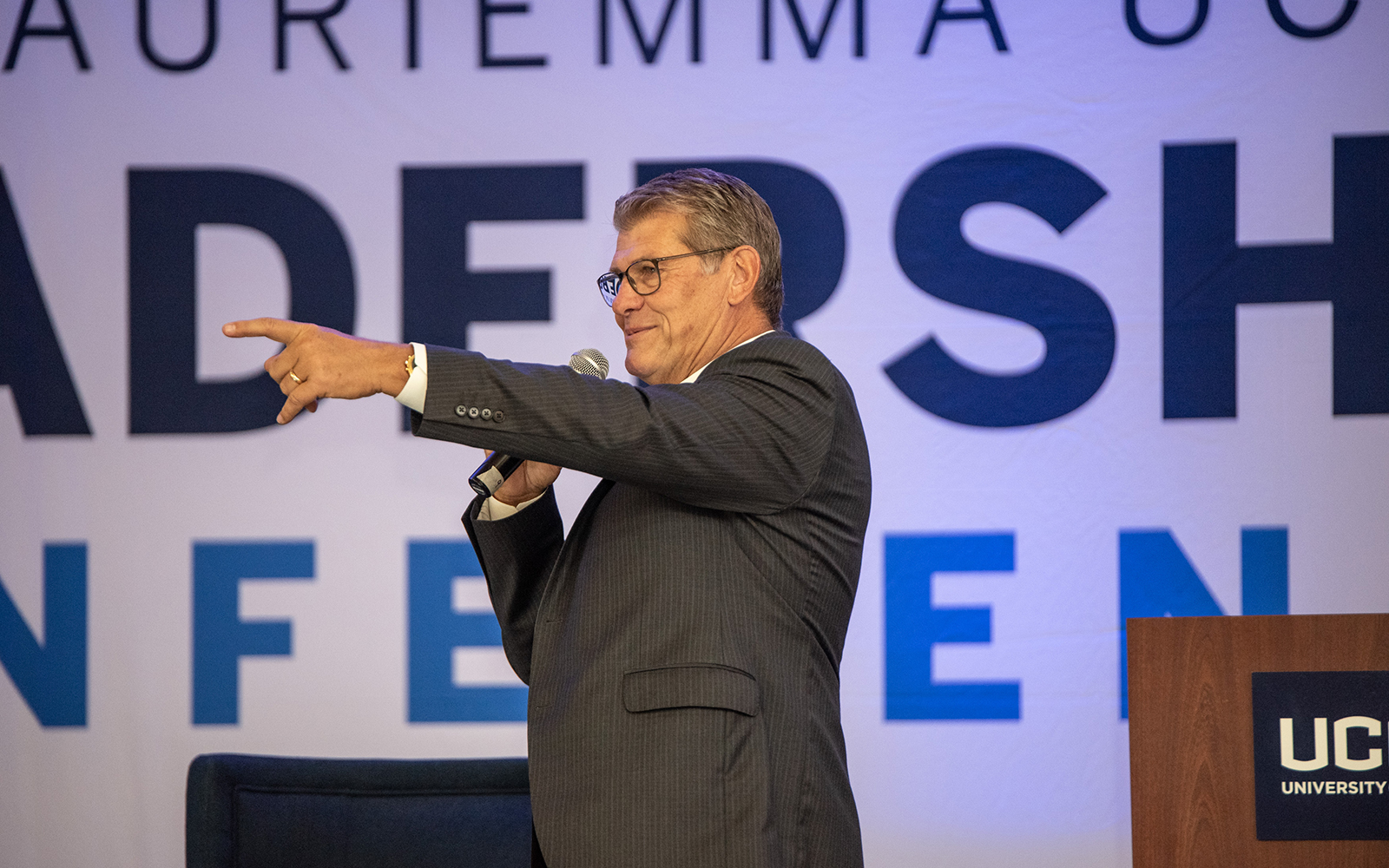 Coach Geno Auriemma stands beside a podium, addressing a crowd.