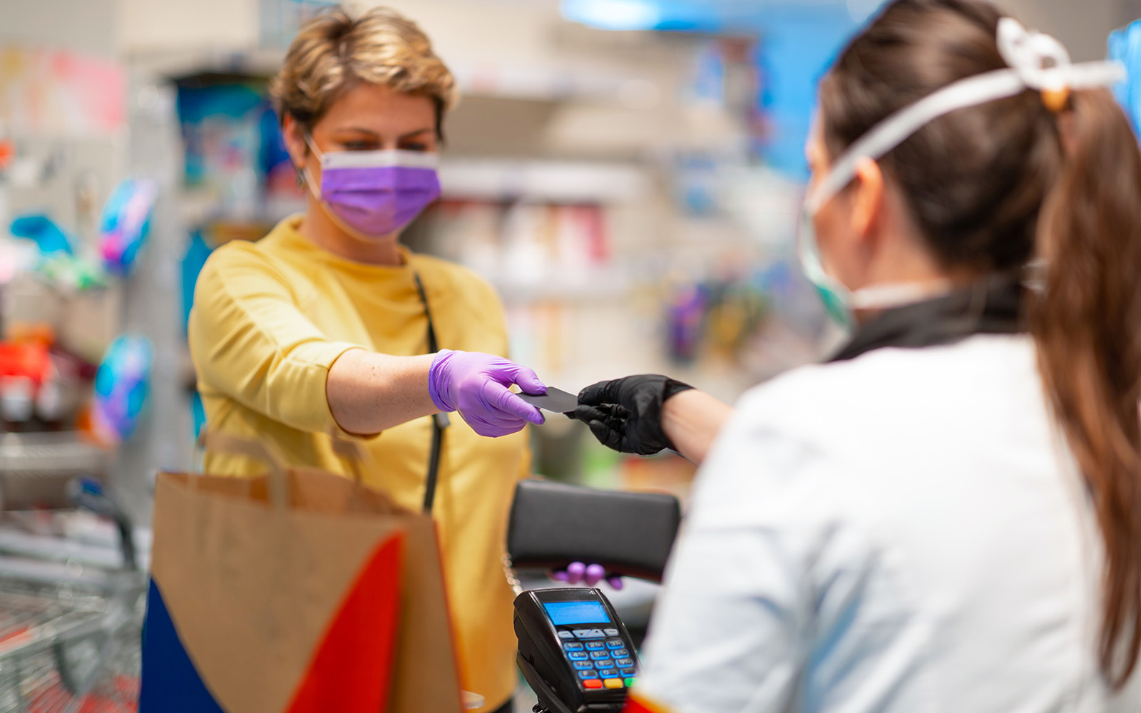 Cashier returning credit card at the cash register to woman with wallet wearing protective face mask and gloves to prevent viruses