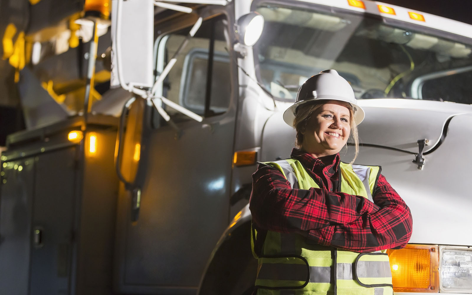 A female worker wearing a hardhat and safety vest standing in front of a work truck. She is a utility worker, engineer or technician.
