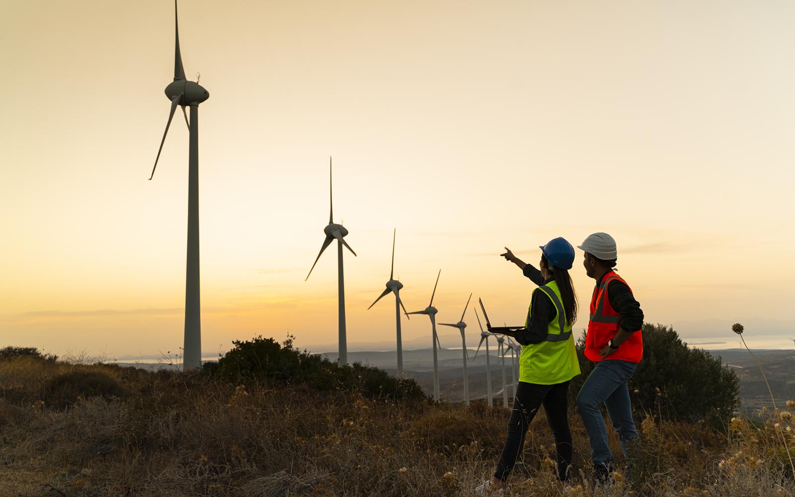 Silhouette of young engineer holding laptop computer planning and working for the energy industry and standing beside a wind turbines farm power station at sunset time